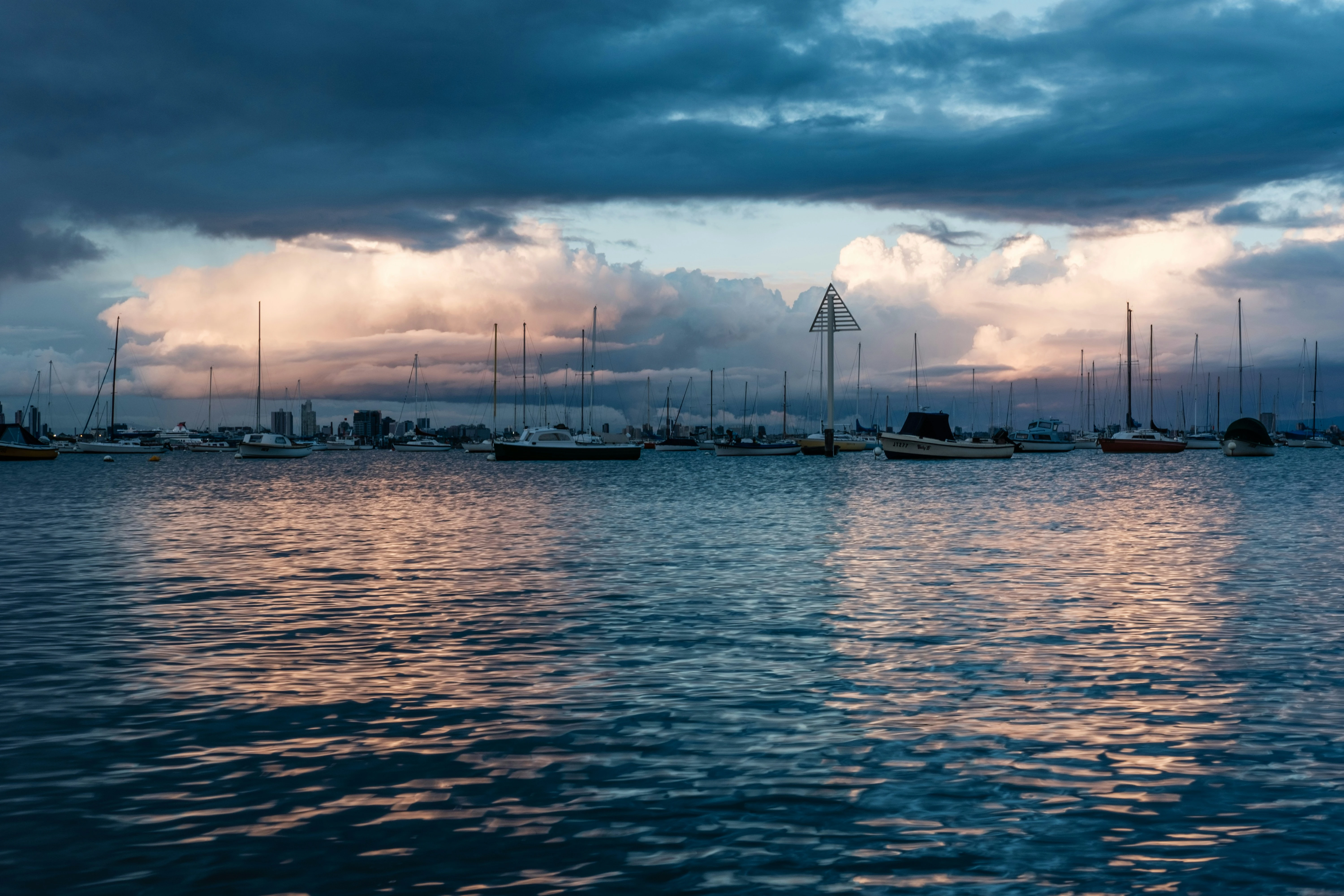 Yachts lying to moorings in a sheltered harbour at dusk, with a bank of storm cloud building over the shoreline behind them