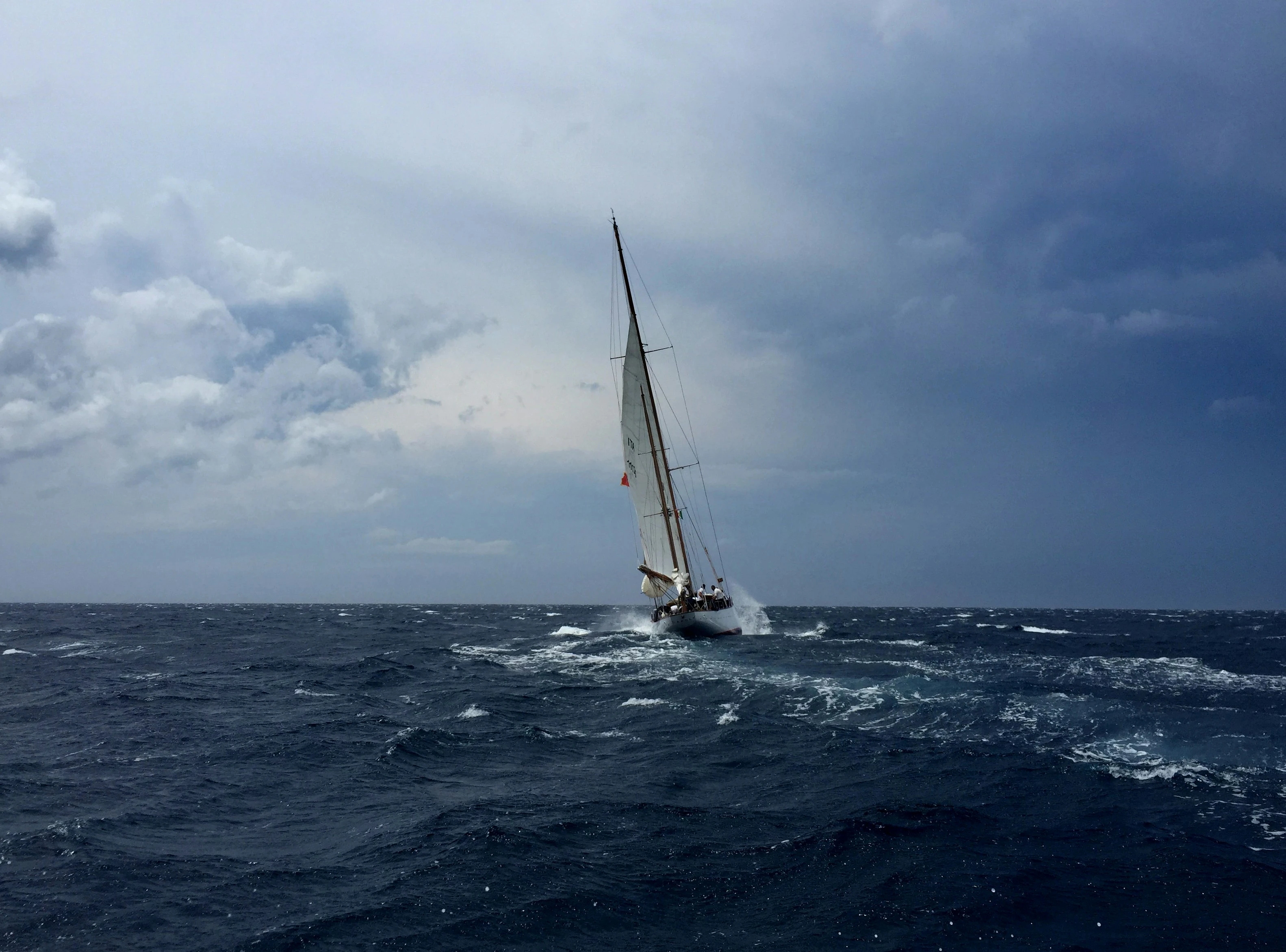 A yacht sailing hard through a big grey sea under a heavy overcast sky, spray coming off the bow